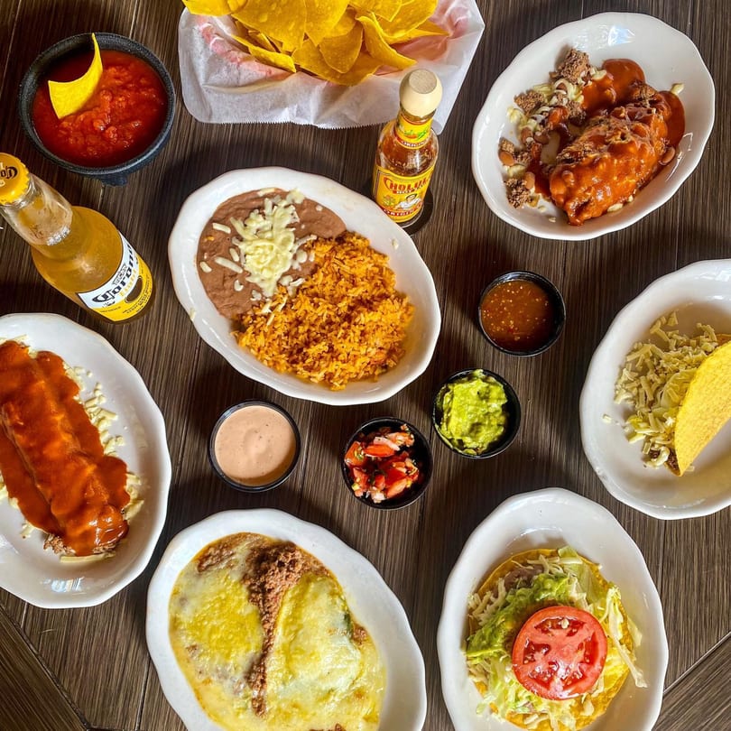 Overhead view of a Mexican feast with enchiladas, tacos, rice, beans, chips, salsa, and beer.