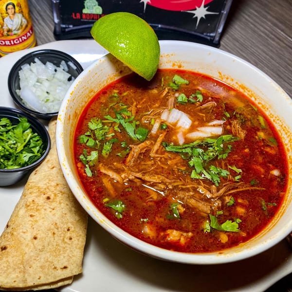 A bowl of Mexican birria soup with shredded meat, cilantro, onions, lime, and tortillas.