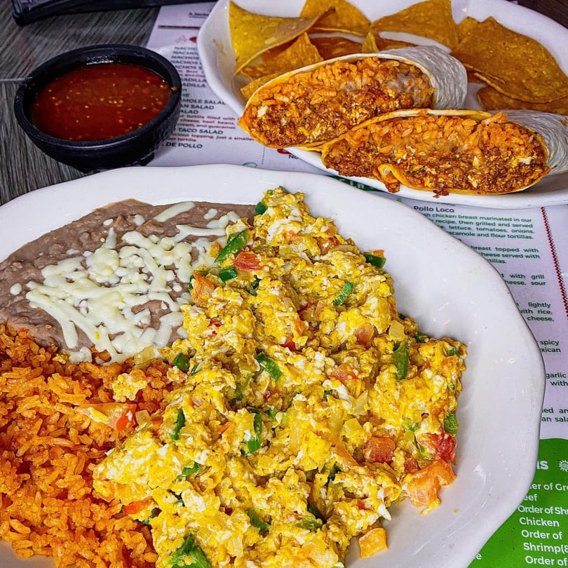 A Mexican breakfast plate with scrambled eggs, refried beans, rice, and two burritos with chips and salsa.